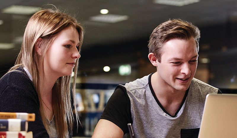 Two students collaborating and working on a laptop in a library setting.