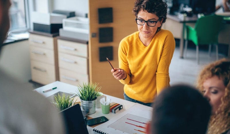 A woman in a yellow shirt leading a discussion with colleagues in an office.
